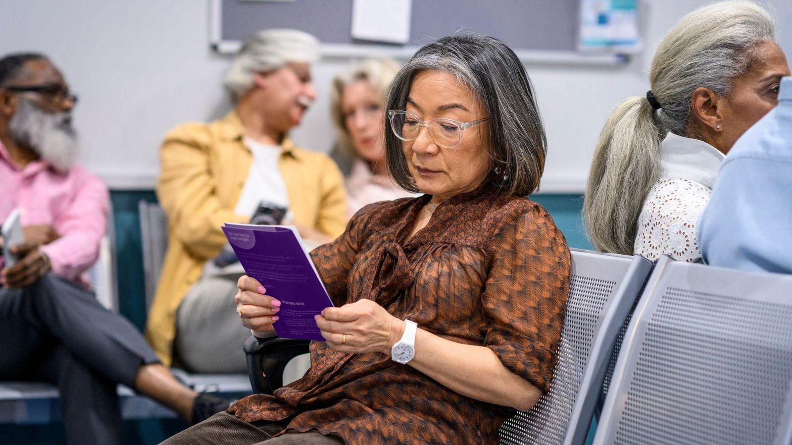 A woman with gray hair sits in a waiting room, reading a purple brochure, while others sit nearby in the background.