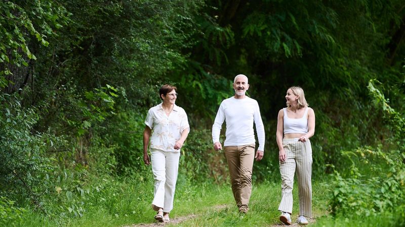 A family of three – Alizée a woman living with type 1 diabetes and both parents - walking along a forest path surrounded by tall trees. They appear to be enjoying a nature walk together
