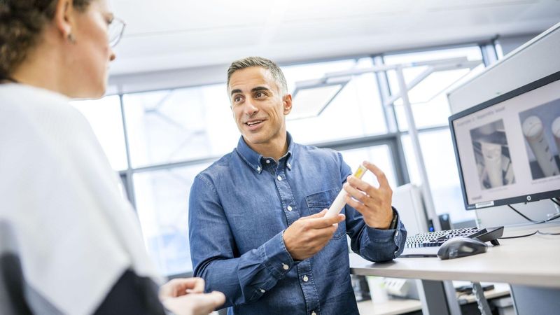 Dr. Nima Aghajari, Lead Data Science & Simulations, Device Central Engineering & Data Science, in a modern office setting holding a cylindrical object while discussing with another person. A computer monitor in the background displays technical diagrams or simulations.