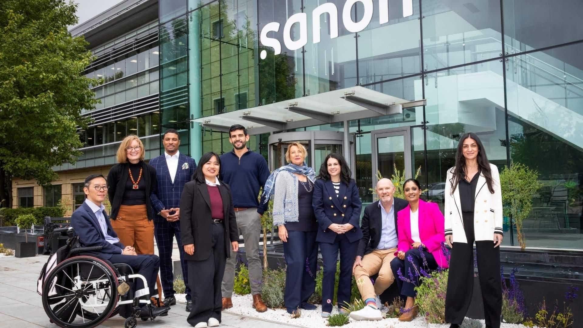 A diverse group of ten professionals in business-casual attire poses for a portrait in front of a modern glass building featuring the Sanofi logo.